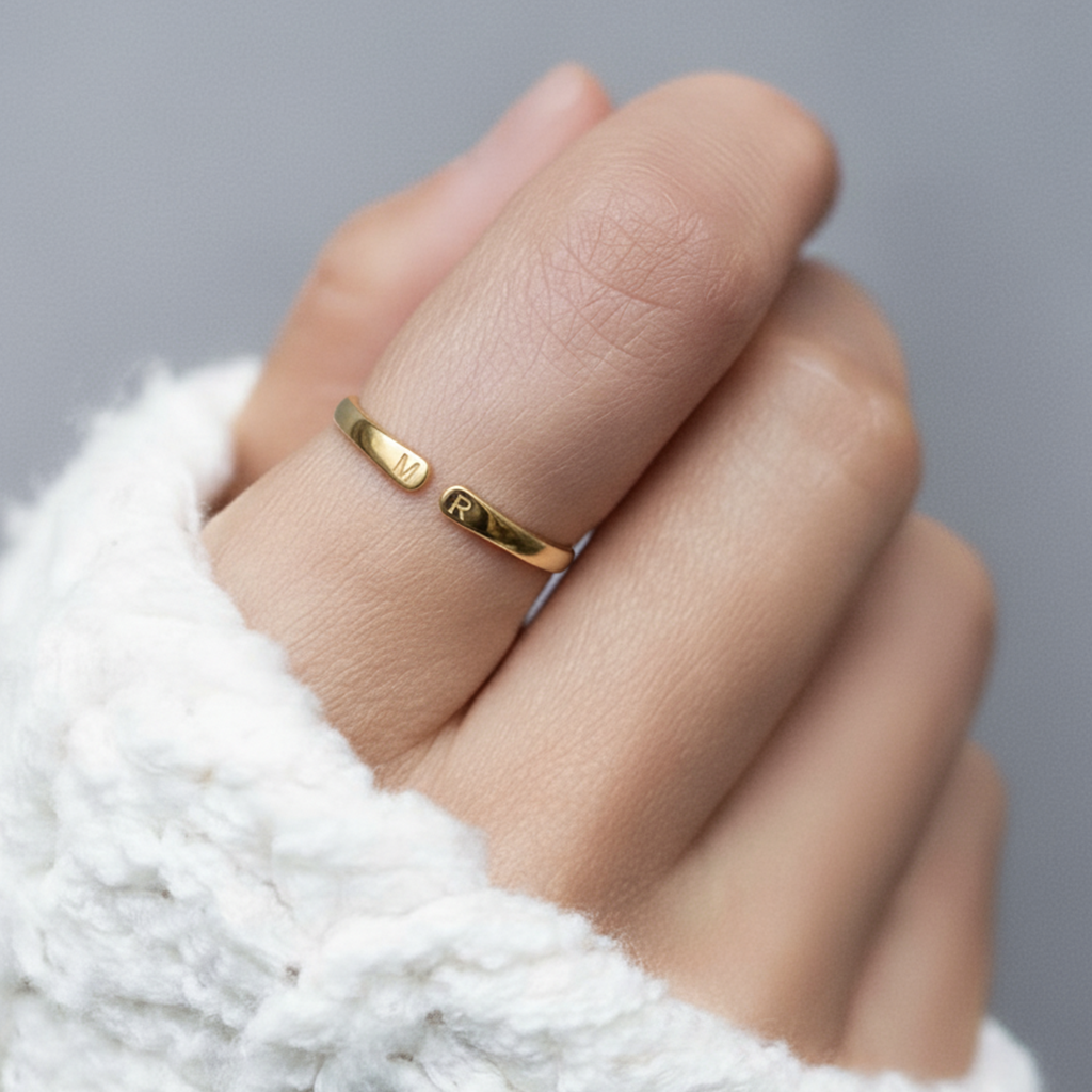 Anillo abierto dorado con iniciales en la mano, sobre un jersey blanco