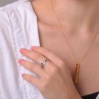 Anillo abrazo plateado en la mano, junto a un colgante de barra dorado grabado con AMAVI