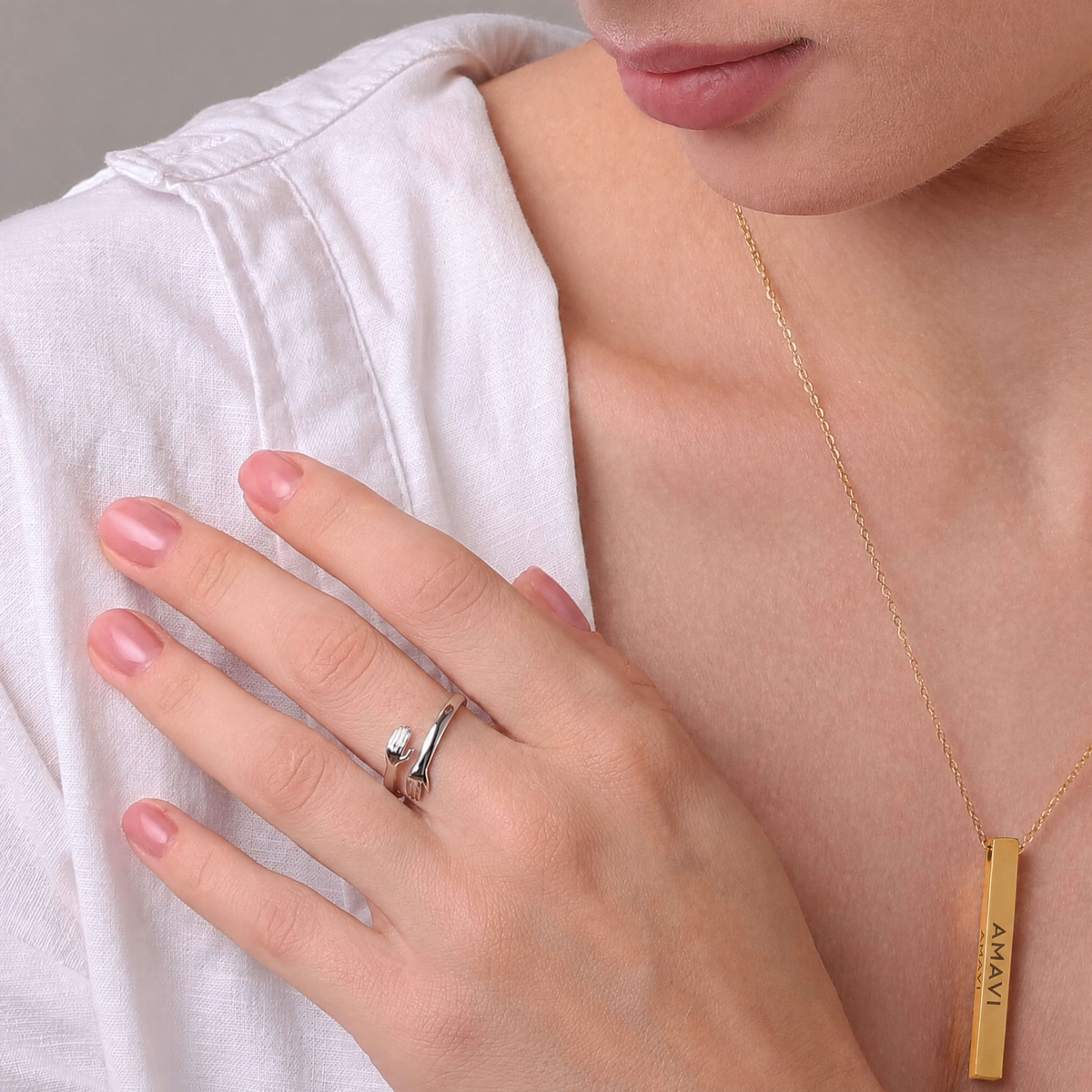 Anillo abrazo plateado en la mano, junto a un colgante de barra dorado grabado con AMAVI